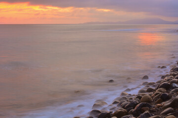 Sunset at sea, calm water surface, large smooth stones on the shore. - 458233055