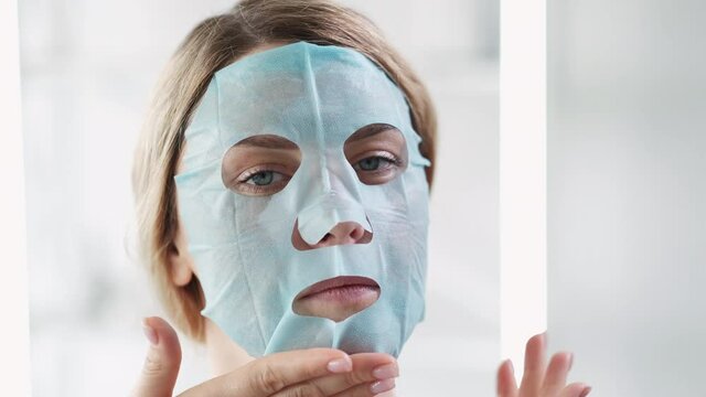 Face mask apply. Skin moisturizing. Facial treatment. Beauty procedure. Closeup portrait of woman rubbing cosmetic anti-aging product in light bathroom interior.
