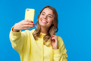 Close-up portrait tender cute blond girl in yellow hoodie, holding mobile phone, taking selfie with smartphone posing adding photo filter in appllication, standing blue background