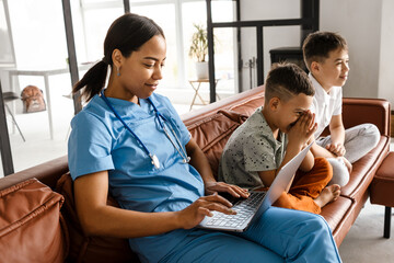 Fototapeta premium Black woman doctor using laptop while sitting with her sons on couch