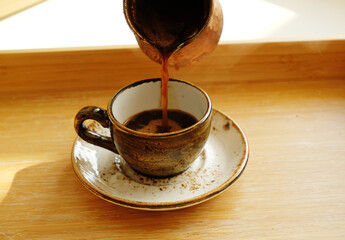 Hot turkish coffee pouring from cezve into ceramic espresso cup on saucer. Bamboo wood tray