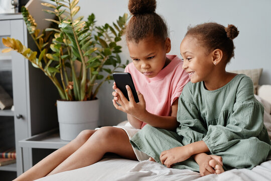 Portrait of two cute sisters using smartphone together while sitting on bed in cozy room, copy space