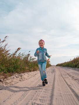 Happy child running jumping having fun on empty autumn sea beach. Blond girl walking on white sand road. Blue sky background. Lifestyle photo. Cheerful kid with bright colorful rainbow pop it toy.