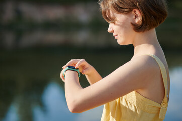 A teenage girl sets up a fitness tracker on the background of the lake. Walking tour.