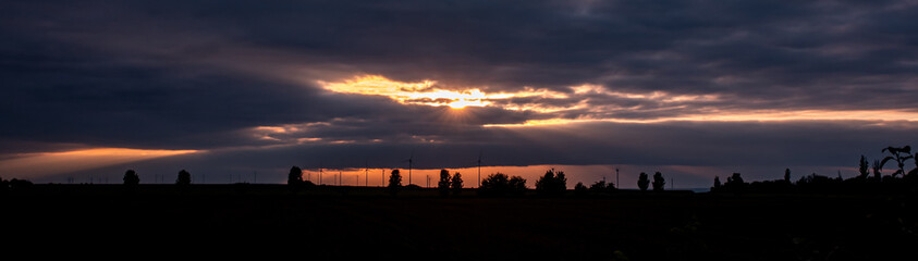 Silhouette of wind turbines and trees at the horizon during sunset. Dramatic orange glowing sky with a lot of clouds, sunbeams shines through the clouds.