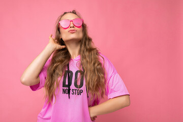 Photo of happy positive cool young beautiful blonde curly woman with sincere emotions wearing stylish pink hip-hop t-shirt and hipster sunglasses isolated on pink background with empty space and