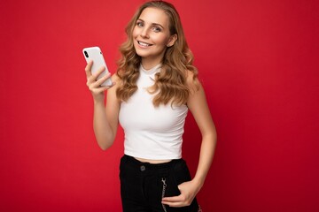 Naklejka premium Smiling happy pretty young blonde woman wearing white t-shirt isolated over red background chatting via mobile phone looking at camera