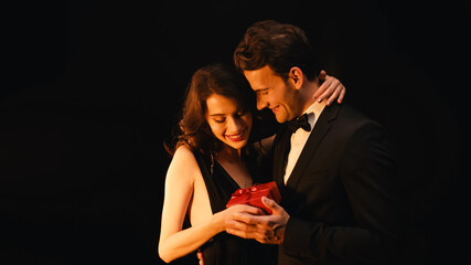 happy young woman receiving gift box from man in suit isolated on black