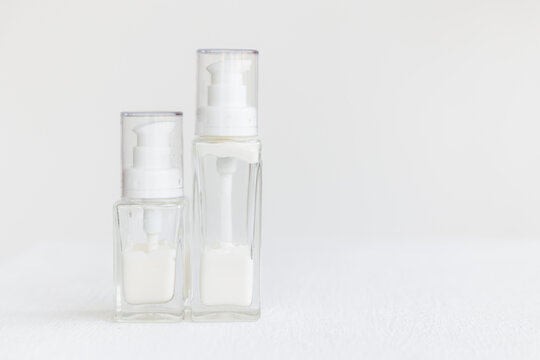 Two Glass Cream Bottles With A Dispenser Lid Stand On A White Cube. White Cream Inside Transparent Containers. Home And Professional Skin Care.