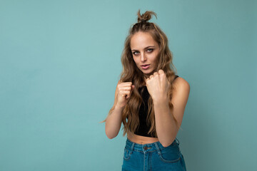 Young beautiful blonde wavy-haired woman with sincere emotions wearing trendy black top isolated over blue background with empty space and ready to fight with fist