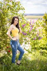 Naklejka na ściany i meble Young woman in yellow top and blue jeans standing in the garden next to a lilac bush in summer