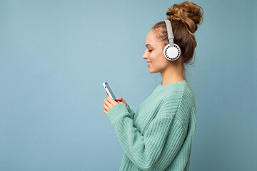 Photo of beautiful joyful smiling young woman wearing stylish casual clothes isolated over background wall holding and using mobile phone wearing white bluetooth headphones listening to music and