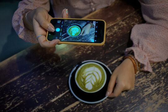 Woman hand holding modern smartphone or cell telephone digital camera taking photo a cup of green tea matcha latte art for post upload file to online on Internet social media resources.