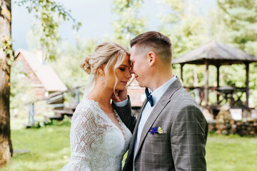 Portrait of young couple in love, newlyweds are flirting with each other, foreheads are touching, groom caresses bride in white dress in park outdoors. Wedding day, marriage concept