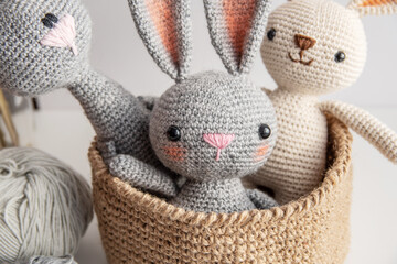 Three handmade bunnies sit in a jute basket on a white table, knitting yarn side by side.