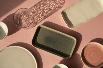 Aesthetic bohemian crystal glass with sparkling sunlight shadow, homemade ceramic plates on the dusty pink background. Flat lay, top view