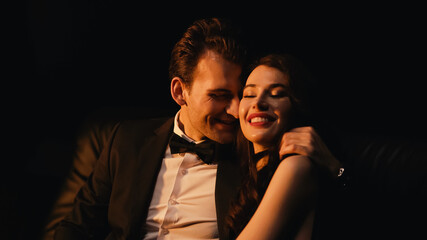 man in suit hugging and flirting with pleased woman isolated on black