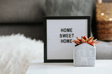 A sign with the inscription home sweet home and a succulent flower in a slide in the shape of a house on a coffee table in the home interior of the living room. Cozy autumn, winter, spring concept.