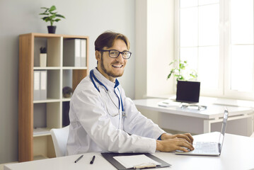 Medical workflow. Portrait of happy young male doctor working on laptop sitting at his workplace in...