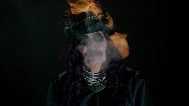 Portrait of sinister senior old mature woman in carnival costume of Halloween witch blows smoke from mouth, trying to scare isolated on black studio background. Horror theme of cosplay clown, vampire