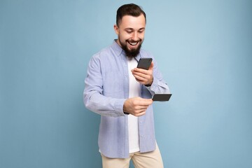 Smiling happy Handsome man wearing everyday clothes isolated on background wall holding and using phone and credit card making payment looking at smartphone screen