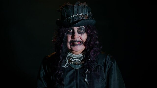 Sinister elderly woman with horrible scary Halloween witch makeup in costume making faces, looking ominous at camera, trying to scare. Portrait of vampire granny isolated against black background