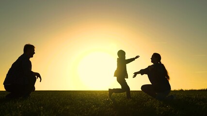 Happy family is playing in park at sunset. Daughter runs from dad to mom in sun. Happy family and childhood concept. Family weekend in the spring on grass. Walk with small kid in nature.