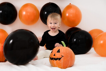 Fototapeta premium Happy blond caucasan infant celebrating Halloween day at home with pumpkin, balloons. Smiling emotional baby girl having fun.Laughing child of 1 year old looking at camera on 31th October holiday