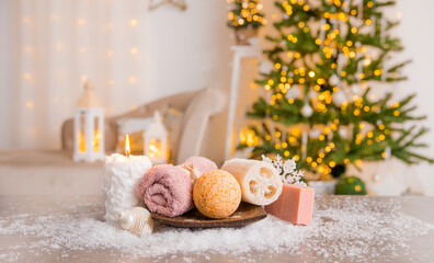 Winter spa pampering concept. Various self care products bath bomb, soap bar, luffa sponge on wood tray with candle burning and Christmas tree with lanterns on background.