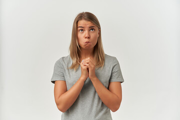 Indoor shot of young adult female, wears gray t-shirt, looks upwards, keeps her palms together in praying position and biting her lips. Isolated over white background