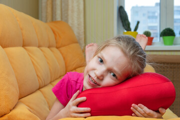 Obraz na płótnie Canvas Funny and cute blonde little laughing girl lying on the red pillow on the orange sofa in the children's room. Children's cheerful emotions. Happy child concept.