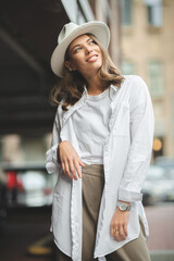 Adorable beautiful young woman expresses happiness, wears fashionable clothes and hat, poses on street, against buildings