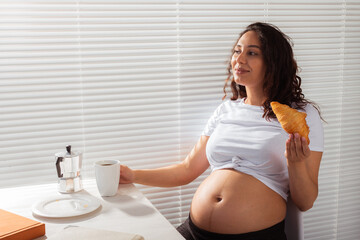 Happy pregnant young beautiful woman eating croissant during morning breakfast. Concept of pleasant morning and positive attitude during pregnancy