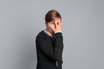 Portrait of shocked and surprised teen boy covering eyes, isolated on grey background, wearing in...