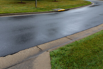 Diagonal view of a rain soaked street surface reflecting gray skies, well maintained green grass...