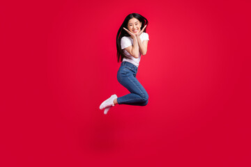Photo of funky adorable korean lady jump hands cheeks wear white t-shirt jeans sneakers isolated red color background