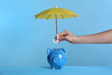 Woman holding small umbrella over piggy bank on light blue background, closeup - Powered by Adobe