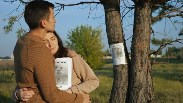 Careful man hugs upset girlfriend in beige hoodie holding papers stack near poster of missing dog stuck on tree trunk in city park at sunset