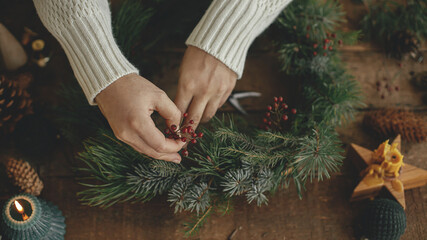 Making rustic christmas wreath close up. Woman hands holding red berries and arranging christmas wreath on rustic wooden background with candle, pine cones, thread. Winter holiday preparations