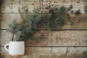 Stylish cup with scattered spruce branches, red berries, pine cones and anise on rustic old wooden background flat lay. Merry Christmas and Happy Holidays! Hello Winter. Space for text