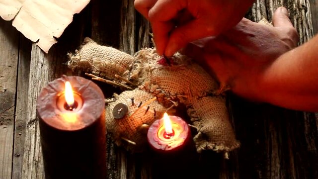 ritual voodoo doll on a wooden table in the light of candles and male hands stabbing her in the heart with a rusty needle