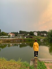 Fototapeta premium A boy in a yellow jacket on the shore of a reservoir. The guy is standing by the lake, view from the back. A teenager on the river bank looks at the water.