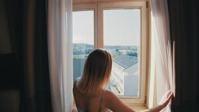Young beautiful blonde woman in elegant sleep lingerie opening dark curtains of window, looking through glass and enjoying cityscape views. Home, wellness, lifestyle.