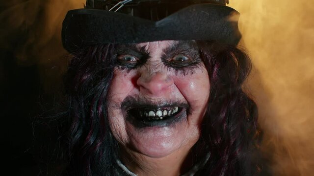Portrait of sinister senior old mature woman in carnival costume of Halloween witch making faces, trying to scare isolated on black studio background. Horror theme of cosplay clown, vampire, beast