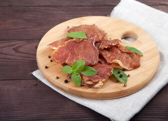Pieces of jerky with basil leaves lie on a wooden cutting board, under it is a linen napkin on a...