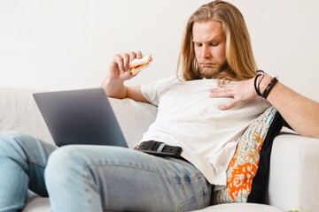 Bearded blonde man eating sandwich while working with laptop