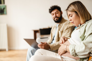 Woman touching her pregnant belly while sitting with her husband on couch