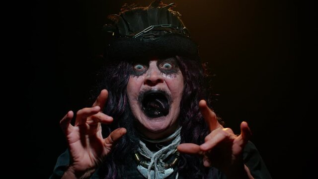 Portrait of sinister senior old mature woman in carnival costume of Halloween witch showing black dirty tongue, trying to scare on black studio background. Horror theme of cosplay clown, vampire beast