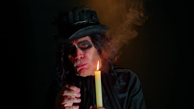 Portrait of sinister senior old mature man in carnival costume of Halloween witcher blows smoke from mouth, making voodoo magic rituals with candle on black studio background. Horror cosplay vampire