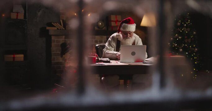 Bearded Santa Claus using laptop, outside view through window, typing on keyboard, buying New Year presents for children online. Grey-haired Saint Nicholas working at night on Christmas Eve. 